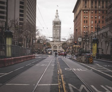 San Franciscos Embarcadero 'nun caddenin sonundaki Ferry Building saat kulesiyle görüntüsü. Tramvay hatları, yayalar ve araçlar şehrin ikonik rıhtım bölgesini bulutlu gökyüzünün altında görüntülemek için dinamik bir sahne oluşturuyor..