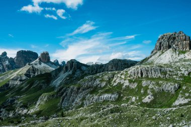 Yeşil vadilerin üzerinden yükselen keskin kayalık tepeleri ve berrak bir yaz gökyüzünün altındaki resimsel Dolomitler manzarası.