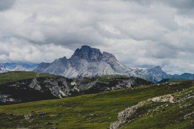 Muhteşem Dolomite zirvesi dramatik fırtına bulutlarının altında. Engebeli kayalık dağ sırtları, ön plandaki yuvarlanan yeşil tepelerle çerçevelenmiş alp manzarasına hakim..