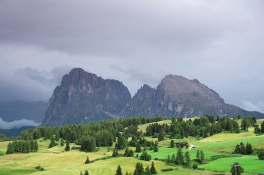 Geniş dağlık alanlar güneş ışığı altında yeşil bir ışık altında parlarken, arka planda sarp kayalıkların tepelerinde dramatik bir karanlık gökyüzü beliriyor..