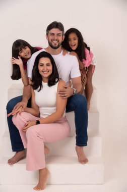 Stüdyoda poz veren neşeli bir aile, bir basamakta oturan ebeveynler ve arka planda iki küçük kızı, gülümseyip oynuyorlar. Atmosfer sevgi ve aile birliği ile dolu..