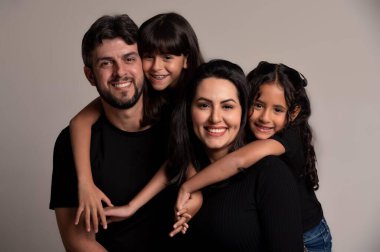 Bir baba, bir anne ve iki kızı gülümserken ve eğlenirken gösteren sevimli bir aile fotoğrafı. Hepsi siyah tişörtler giyer. Tarafsız bir zemine karşı sevgi ve birlik gösterirler. Neşeli ifadelerini ve güçlü duygusal bağlarını vurgularlar..