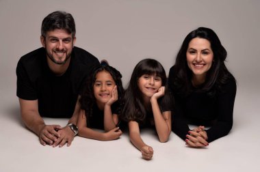 Neşeli bir aile fotoğraf stüdyosunda poz veriyor. Anne ve baba, her ikisi de siyah giyinmiş, iki kızlarına yakın yatıyorlar. Kızlar neşeli gülücükler, gevşek saçlar ve mutlu ifadeler sergiliyorlar. Tarafsız arkaplan onların sevgi ilişkilerini vurgular,