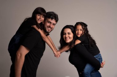 Gülümseyen bir ailenin çekici bir görüntüsü. İçinde bir baba, bir anne ve iki kız var. Koyu renk kıyafetler giyiyorlar, sevgi ve birlik gösteriyorlar. Çocuklar ailelerinin sırtında, eğlence ve sevgiyi sembolize ediyorlar, hepsi tarafsız bir arkaplanda poz veriyorlar.