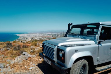 Yunanistan 'ın Girit kentindeki Heraklion' a bakan bir tepede gümüş bir araç park halinde. Araba bir Jeep Wrangler ve toprak bir yolda park halinde. Arabadan manzara okyanusu ve aşağıdaki şehri gösteriyor. Huzur dolu bir sahne.
