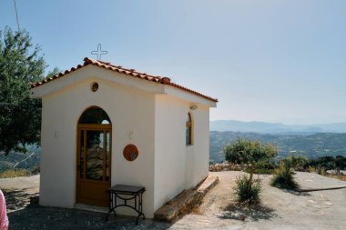 Kırmızı çatılı küçük beyaz bir bina bir tepenin üzerinde duruyor. Binanın bir penceresi ve kapısı var. Tepe ağaçlar ve çalılarla kaplı.