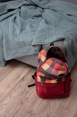 Odadaki yatağın yanında yerde duran kırmızı renkli kareli sırt çantası. Temizlik ve disiplin.