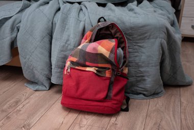 Odadaki yatağın yanında yerde duran kırmızı renkli kareli sırt çantası. Temizlik ve disiplin.
