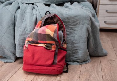 Odadaki yatağın yanında yerde duran kırmızı renkli kareli sırt çantası. Temizlik ve disiplin.