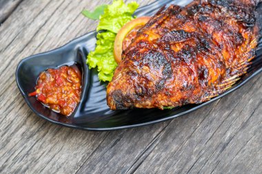 Izgara balık (ikan bakar) siyah bir tabak üzerinde sambal, marul ve domates ile yakın plan, kırsal ahşap bir masa karşı kurulmuş.