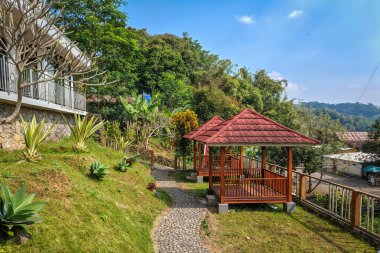 Huzurlu bir tepe inzivası. Üzerinde iki ahşap çardak ve kırmızı çatılar var. Taş döşeli bir yol ve açık mavi gökyüzünün altında yemyeşil bir yer..