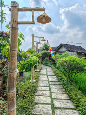 Bambu lambaları ve canlı yeşilliklerle kaplı, bereketli bir bahçe boyunca uzanan, taşlarla döşenmiş sakin bir yol bulutlu bir gökyüzünün altındaki geleneksel bir eve doğru esiyor..