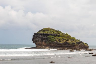 Sahil manzarası, kumlu bir sahil, yumuşak dalgalar ve açık mavi gökyüzünün altında birkaç kayalık ada içerir..