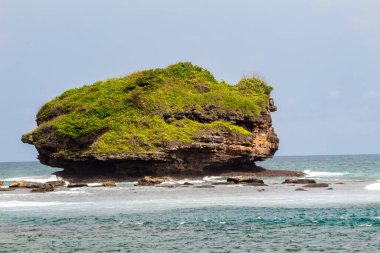 Sahil manzarası, kumlu bir sahil, yumuşak dalgalar ve açık mavi gökyüzünün altında birkaç kayalık ada içerir..