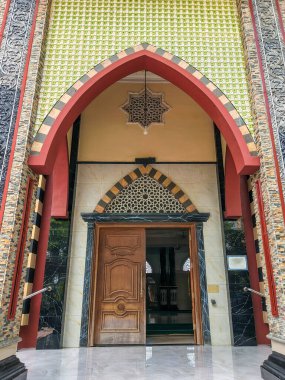 The vibrant, ornate entrance and arched doorway of a mosque, featuring colorful geometric tiles, intricate carvings, and a large wooden door, showcasing Islamic architecture.