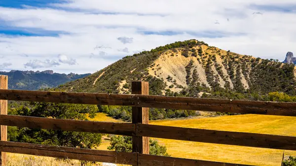 Ön plan boyunca uzanan tahta bir çit, çarpıcı erozyon çizgileri ve Colorado kırsalında dağınık bir çalılıkla güneşli bir yamacı çerçeveliyor. Arkasında, San Juan Dağları yumuşak, bulutlu bir gökyüzünün altında hafifçe yükselir..