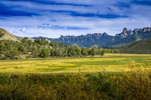 Altın çayırların ve dağınık ağaçların geniş bir manzarası Colorado 'daki sivri bir dağ sırasının altında ortaya çıkar. Dramatik kaya oluşumları ve katmanlı tepeler derin mavi, bulut çizgili bir gökyüzü karşısında klasik bir Amerikan Batı manzarası yaratıyor..