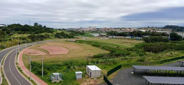 Panoramik hava görüntüsü, yemyeşil bitki örtüsü ve dolambaçlı bir yolu olan bir şehir parkını gösteriyor. Arka plandaki şehirle zıt yönde, bulutlu bir gökyüzünün altında binalar ve altyapılarla dolu..