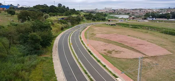 Kentsel bir alanda, çalı ve ağaçlarla dolu yeşil bir alanda kıvrılan dolambaçlı bir yolun hava görüntüsü. Arka planda, konut binaları ve bulutlu bir gökyüzü olan bir şehir karşıtlık yaratır.