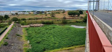 Bir köprüden görülen canlı yeşil bir arazinin geniş bir görüntüsü. Sahne, çevreleyen bitki örtüsüyle çelişen, gökyüzünde karanlık ve aydınlık bulutları içerir. Arka planda, kentsel binalar doğal alanlarla harmanlanarak sakin ve davetkar bir şehir yaratıyor.