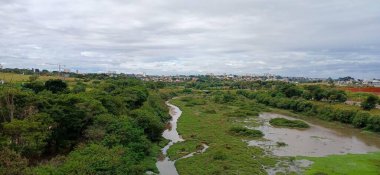 Güzel bir kırsal alan, yemyeşil çayırlarda akan dolambaçlı bir nehri gösteriyor. Yoğun ağaçlar ve yeniden ağaçlandırılmış bitki örtüsü nehir kıyıları boyunca uzanır ve arka planda bir şehir görülebilir. Bulutlu gökyüzü sakin bir atmosferi yansıtıyor..