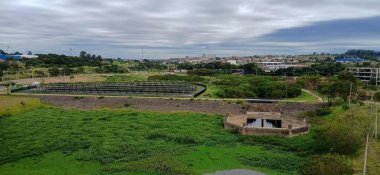 Arka planda modern bir şehirle çevrili, solunda tesisleri olan yemyeşil bir bölgenin panoramik görüntüsü. Bulutlu gökyüzü serin bir atmosferi yansıtırken, bulut gölgeleri yeşil alanlarla güzel bir tezat oluşturuyor. Ortalık sakin, evokin.