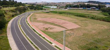 Arkaplanda binalarla çelişen, tarlalar ve yeşilliklerle çevrili, gelişmekte olan bir kentsel bölgedeki dolambaçlı bir yolun hava görüntüsü. Yarı bulutlu hava durgunluğu ve büyümeyi çağrıştırıyor.