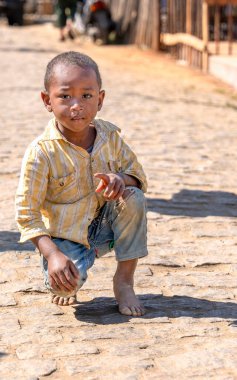Çizgili sarı tişörtlü ve yırtık kotlu çıplak ayaklı genç bir Malagasy köyün parke taşı sokağında çömelmiş. Doğal duruşu ve tuhaf ifadesi, ılık toprak tonlarıyla birleşince Madagaskar 'daki kırsal yaşamı resmediyor..