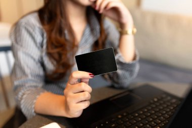 İş elbiseli genç bir kadın dizüstü bilgisayarının yanında boş bir siyah kart tutuyor. İnternetten alışveriş, e-ödeme ya da kimlik güvenliğini simgeliyor. Kapalı alan, yumuşak odaklı arka plan, modern çalışma alanı