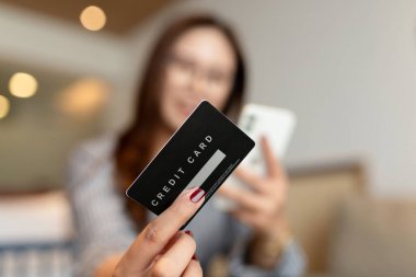 Akıllı telefonunu kullanırken elinde siyah bir kredi kartı olan bir kadının çevrimiçi alışveriş ya da dijital ödeme önerisi. Arka plan, yumuşak ışıklandırmalı, modern bir yatak odasını gösteriyor.