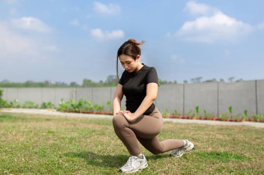 Aktif giyimde odaklanmış bir kadın güneşli gökyüzünün altındaki çimenli alanda derin hamleler yapıyor.