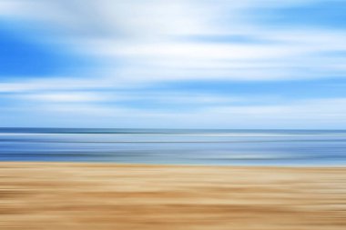 Hareket bulanıklığı olan soyut plaj sahnesi, kumları, sakin deniz dalgalarını ve parlak mavi gökyüzünü gösteriyor..