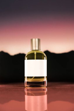 Parlak beyaz etiketli minimalist bir parfüm şişesi dramatik bir gün batımı gradyanı ve dağ siluetine karşı durarak zarafet ve doğal cazibeyi çağrıştırıyor..