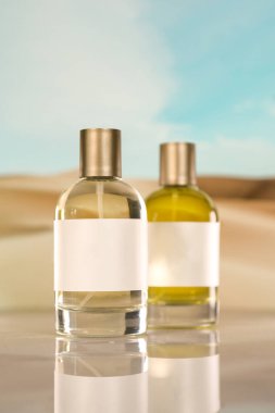 Boş beyaz etiketli iki parlak parfüm şişesi yumuşak bir çöl manzarasına karşı yansıtıcı bir yüzeyde duruyor. Lüks, saflık ve doğal bir estetiği çağrıştırıyor..