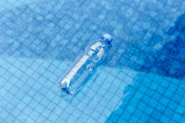 Plastik bir su şişesi yüzme havuzunun yüzeyinde yüzer. Altında mavi fayanslar görülür. Bu da kirliliği ve atığı sembolize eder..