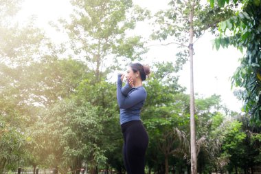 Spor kıyafetli bir kadın açık havada yemyeşil bir parkta ısınırken kollarını göğsüne uzatıyor.
