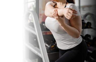 Spor salonunda kolunu uzatan, beyaz tişört ve siyah tayt giyen bir kadının yakın çekimi. Arka planda dambıllar ile fitness ayarları ısınma ve egzersiz hazırlıklarını vurguluyor