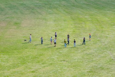 Bir grup çocuk güneş ışığında büyük yeşil bir alanda oynuyorlar.