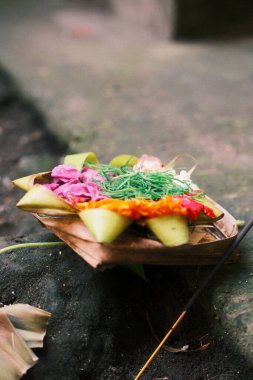 Yakın çekimde geleneksel bir Balili Canang Sari sunumu, canlı renkli çiçekler ve tütsü çubuğuyla yeşil yapraklarla örülmüş küçük bir palmiye yaprağı sepeti yer alıyor.