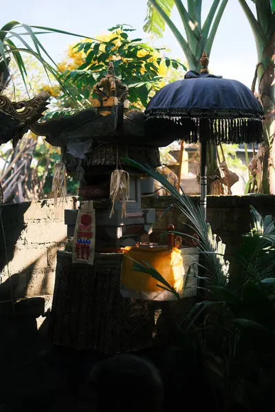 Geleneksel bir Bali taşı sunağı, adaklar ve kumaşlarla süslenmiş, yemyeşil tropikal yaprakların arasında duruyor, koyu mavi bir tören şemsiyesi sahneye ekleniyor.