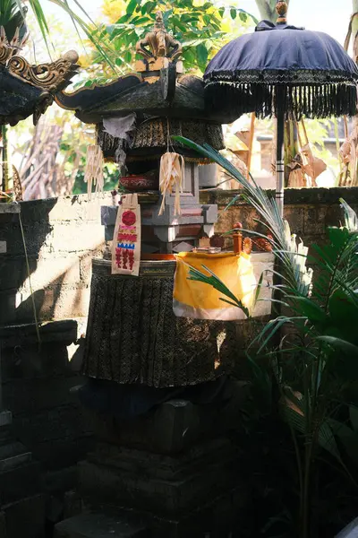 Geleneksel bir Bali taşı sunağı, adaklar ve kumaşlarla süslenmiş, yemyeşil tropikal yaprakların arasında duruyor, koyu mavi bir tören şemsiyesi sahneye ekleniyor.