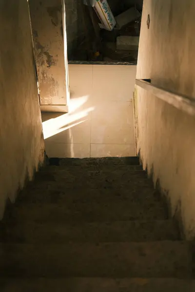 Yukarıdan bakıldığında, aşınmış ve kirli beton bir merdiveni aşağı doğru çeker, güneş ışığı alttaki fayanslı zeminde yaprak gölgeleri bırakır.