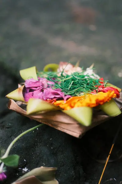 Yakın çekimde geleneksel bir Balili Canang Sari sunumu, canlı renkli çiçekler ve tütsü çubuğuyla yeşil yapraklarla örülmüş küçük bir palmiye yaprağı sepeti yer alıyor.