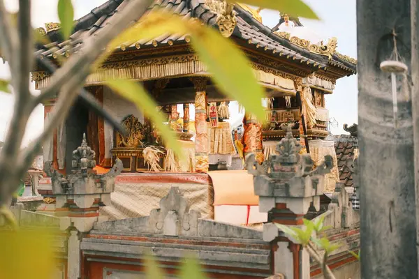 Karmaşık altın ve kırmızı oymalarla süslenmiş geleneksel bir Bali tapınağı, kısmen bulanık yeşil yapraklar ve ön planda bir taş sütun tarafından çerçevelenmiştir.