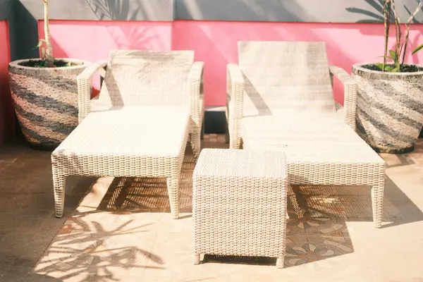 İki açık renkli hasır (rattan) şezlong salonu ve uyumlu bir yan masa canlı pembe ve gri bir duvara karşı döşenmiş fayanslı bir veranda üzerine dizilmiştir.
