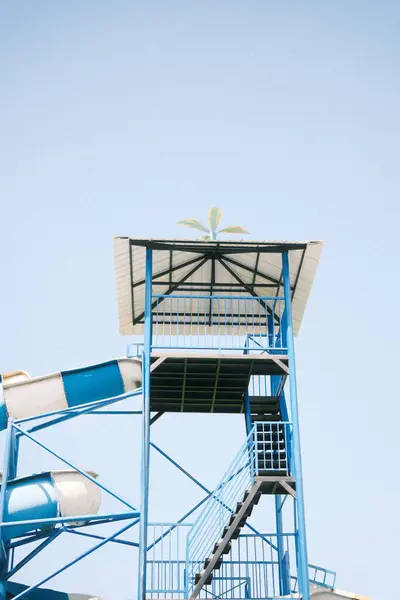 Su kaydırağı kulesinin üst kısmının temiz bir görüntüsü, mavi metal çerçeve, merdiven ve parlak, berrak bir gökyüzüne ayarlanmış bir kaydırağın tepesi.