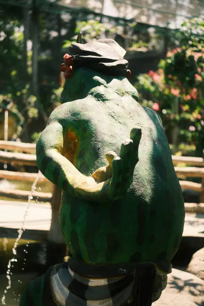 Kaprisli yeşil bir kurbağa heykeli, muhtemelen çimento veya taştan yapılmış, bir bahçe ortamında duruyor, arkasında hafifçe su sıçrıyor.