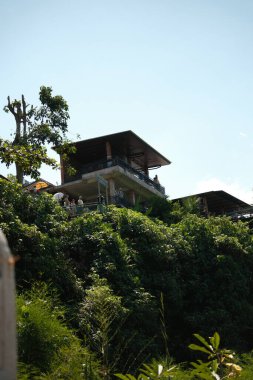 Tegenungan Şelalesi, Bali - 27 Nisan 2025: Balkonları ve kendine özgü çatı çizgisi olan modern çok katlı bir bina, sarp, yemyeşil bir yamaçla güzelce bütünleşmiştir.