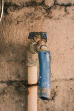 Eski, çürümüş, mavi saplı, yıpranmış bir musluğun yakın çekimi, eskimiş borulara yapışmış, çürüme ve kullanım temaları vurgulanıyor.