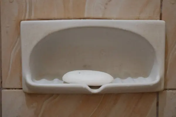 Beyaz ya da beyaz seramik bir sabun kabının yakın plan görüntüsü fayanslı bir duvara monte edilmiş, içinde tek bir kalıp beyaz sabun var.